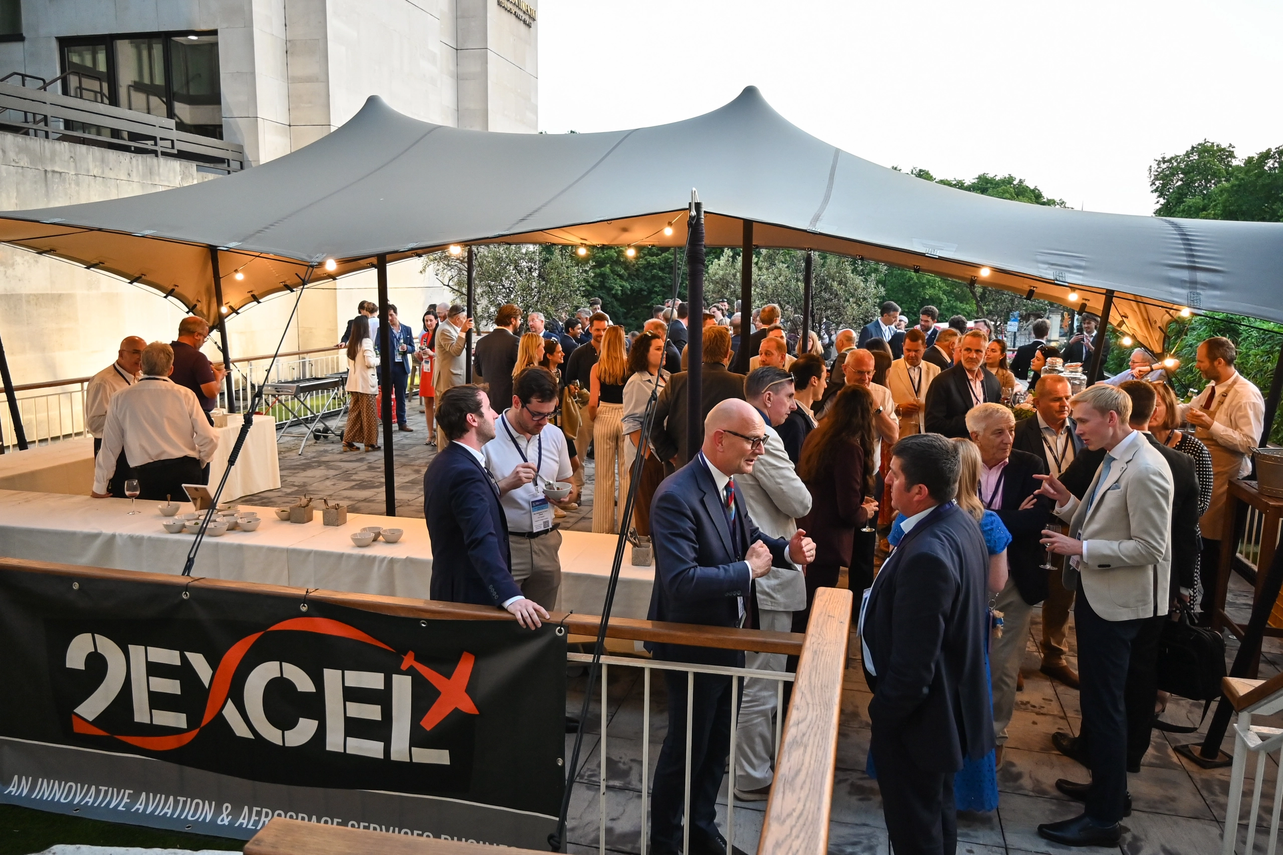 Professional crowd mingling at an outdoor reception under a curved white canopy; banner with 'EXCEL' visible in the foreground.