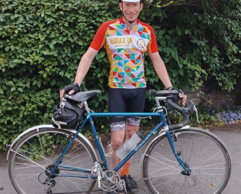 Cyclist in a colorful geometric jersey and white helmet standing beside a blue road bike on a paved path with green hedges in the background.
