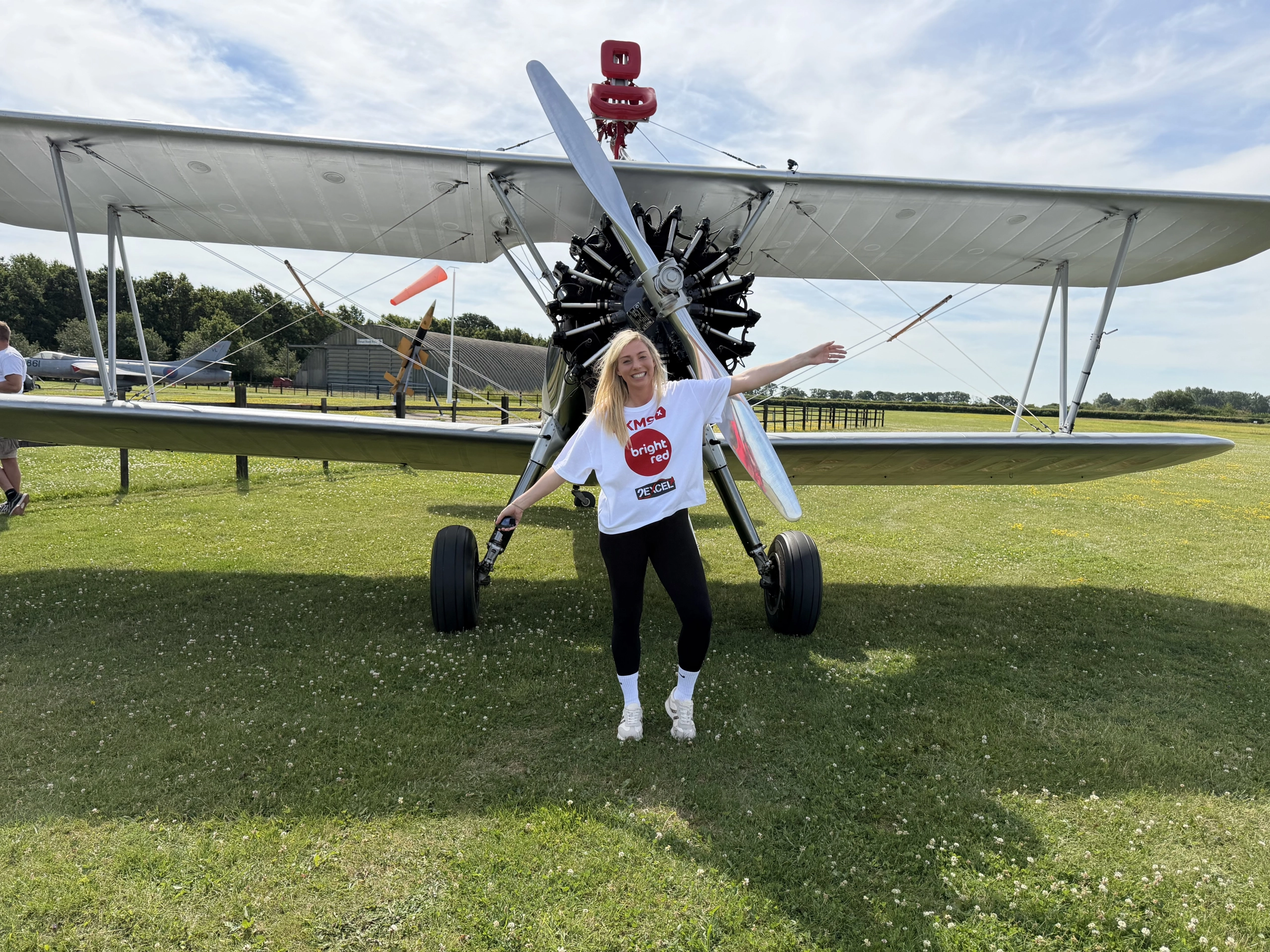 blonde woman in front of bi-plane making aeroplane wings with her arms