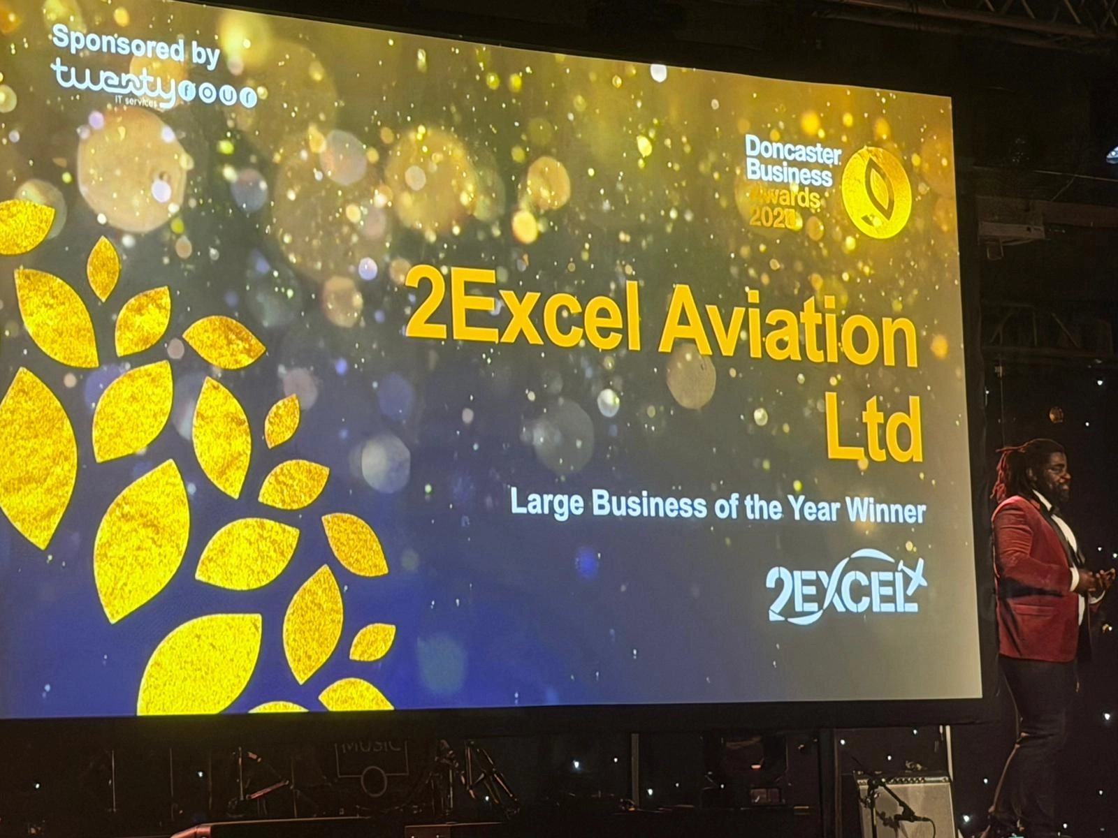 Large screen showing award text: '2Excel Aviation Ltd' as 'Large Business of the Year Winner' at a ceremony, with gold decorative leaves and sponsor logos.